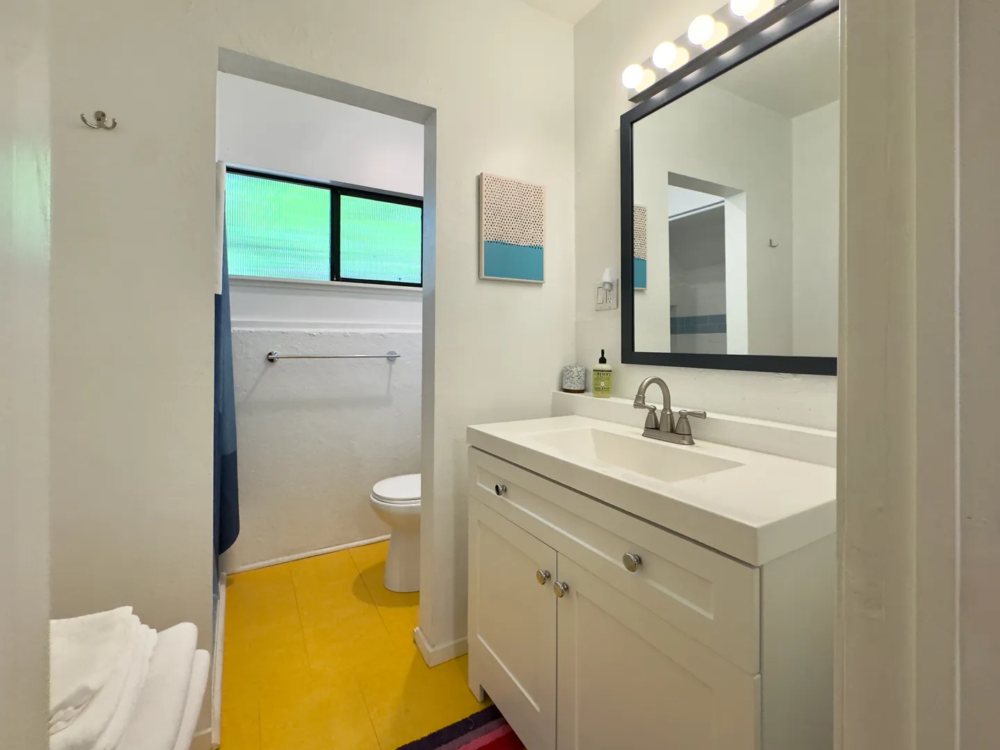 Bathroom with white vanity, mirror with vanity lights, yellow flooring, and an open doorway leading to a toilet area.
