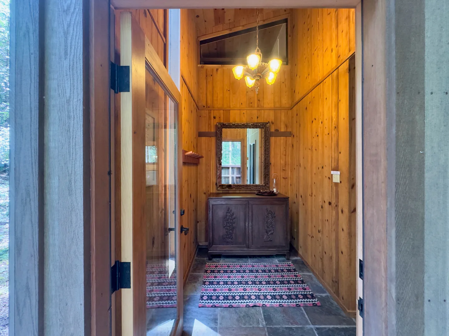 Entryway with vertical wood-paneled walls, stone tile floor, patterned rug, wooden console cabinet, and an ornate mirror beneath a hanging light fixture.