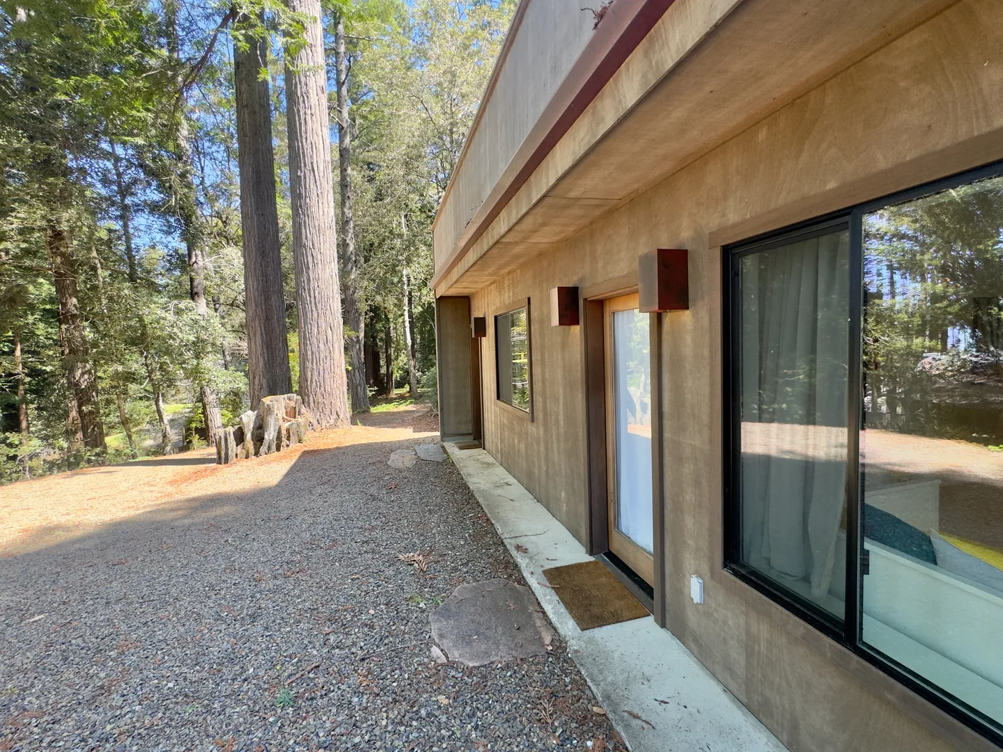 Exterior side view of a brown home with a gravel area alongside tall trees.