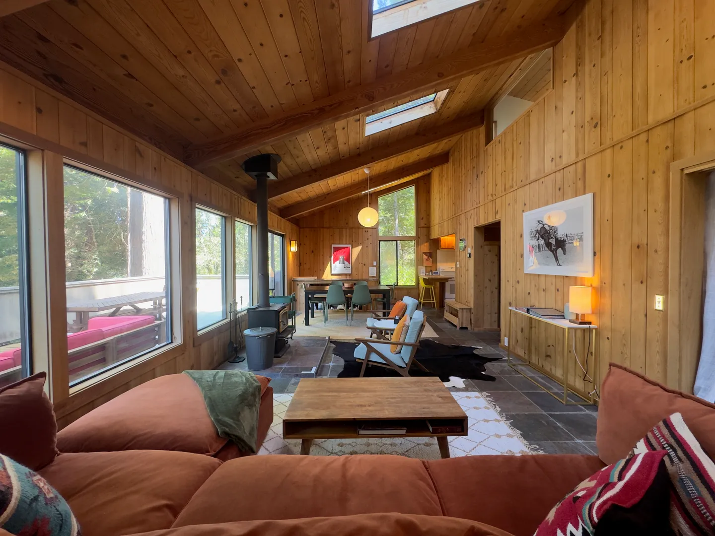 Open-concept living room and dining area featuring wood-paneled walls and ceiling, a wood-burning stove, large windows, and a plush sectional sofa.