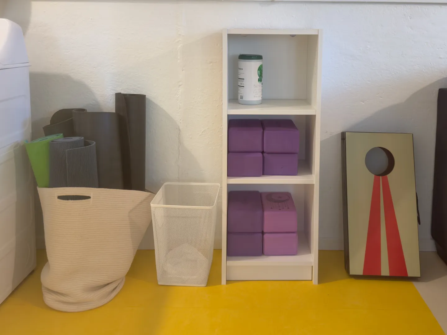 A fitness and game storage area featuring yoga mats in a fabric bin, foam yoga blocks in a white shelving unit, and a cornhole board against a wall.