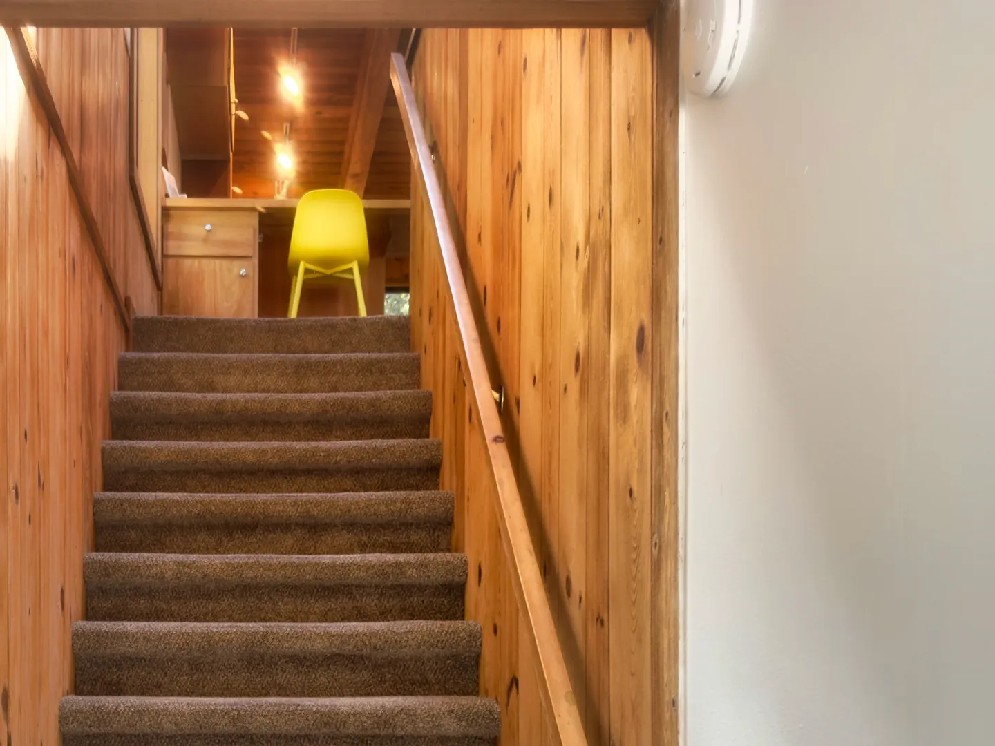 A straight flight of carpeted stairs leading up between wood-paneled walls toward an upper-level room with a yellow chair.