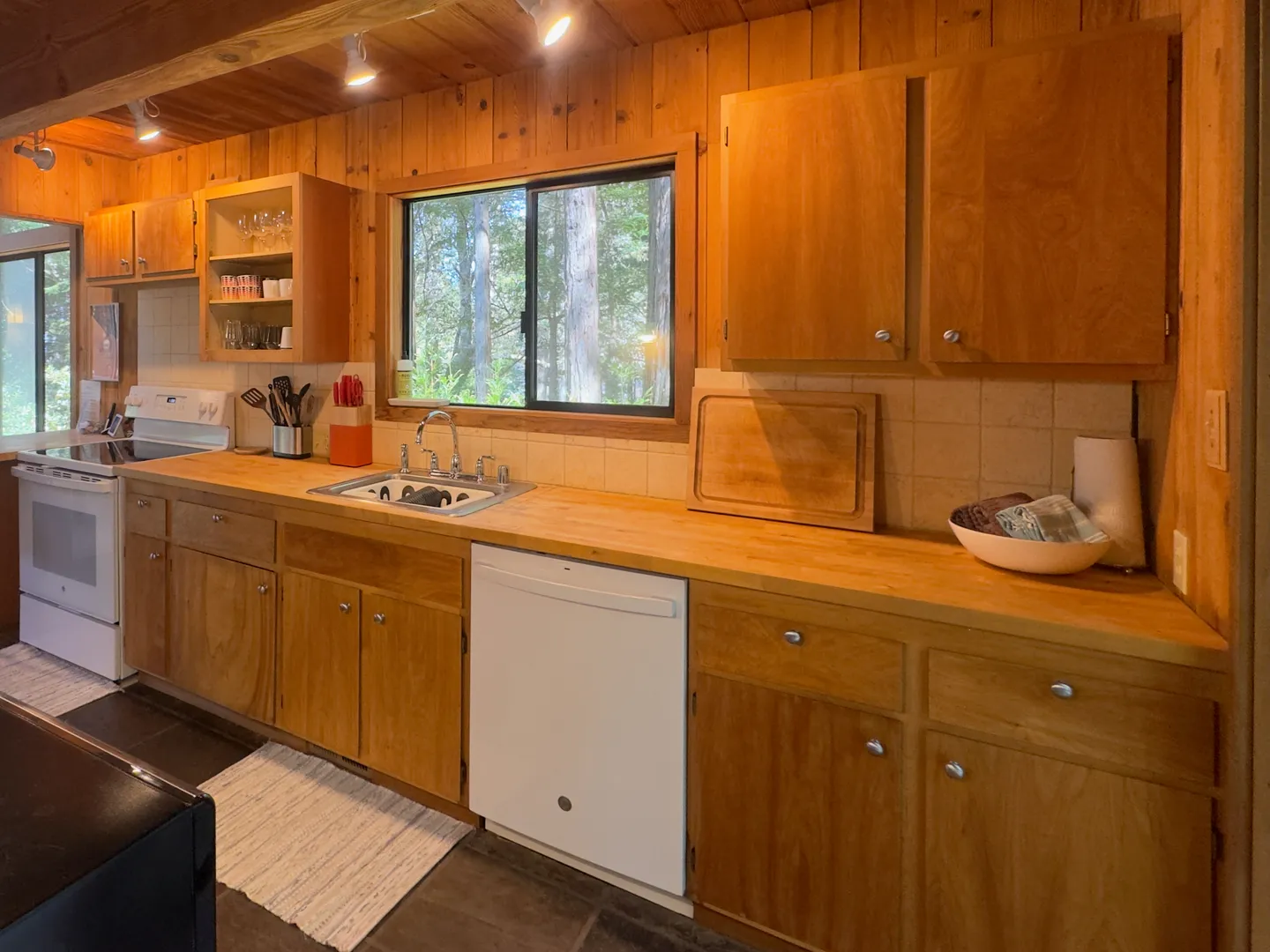 Kitchen featuring wood paneling, wooden cabinets, a butcher block style countertop, a white dishwasher, and a window above the sink.