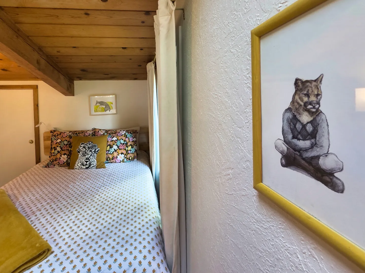 Bedroom nook with a wood-plank ceiling, dressed bed, framed artwork, and textured white walls.