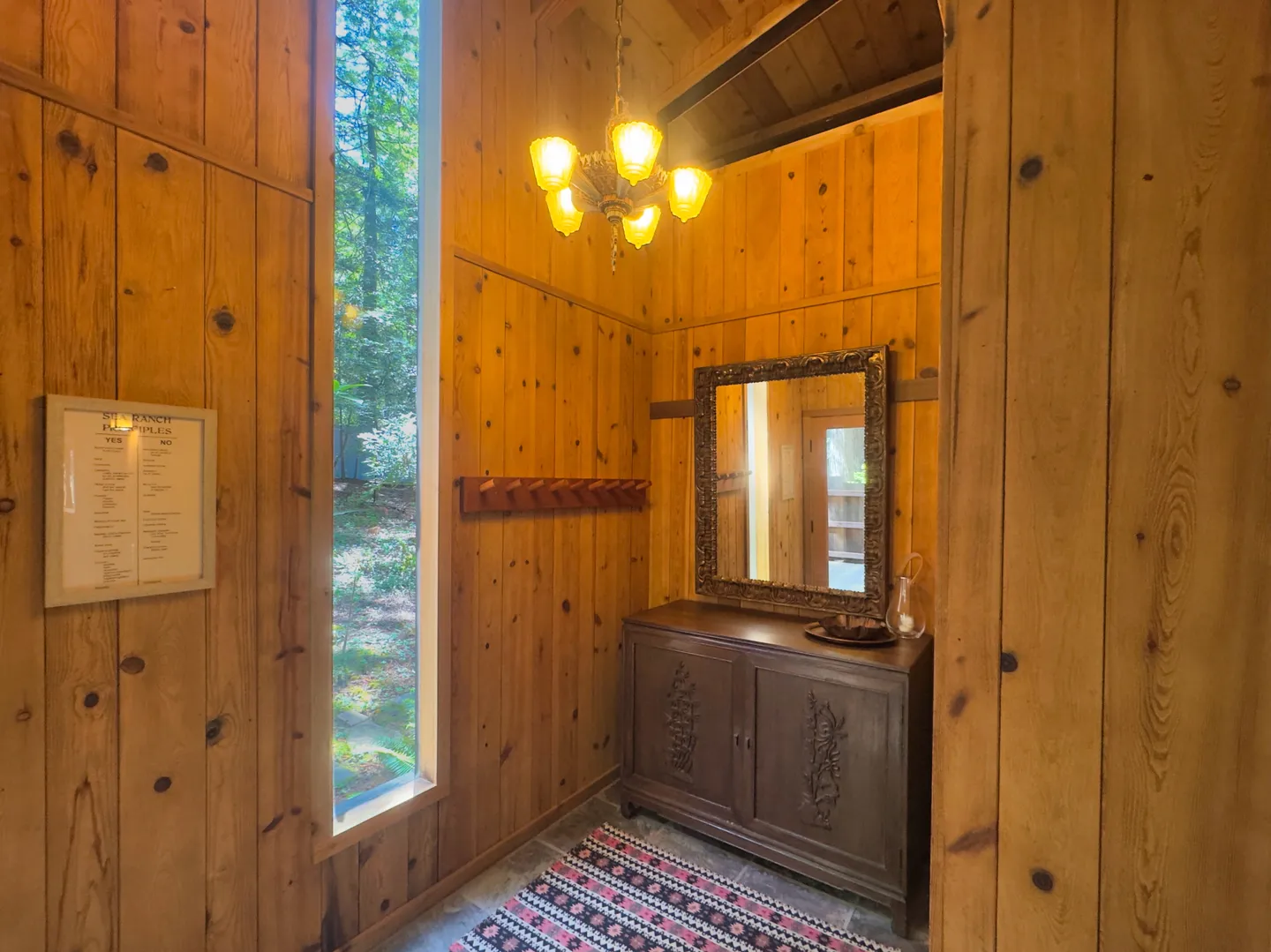 Wood-paneled entryway with a sideboard, decorative mirror, coat rack, and tall vertical window looking out to trees.