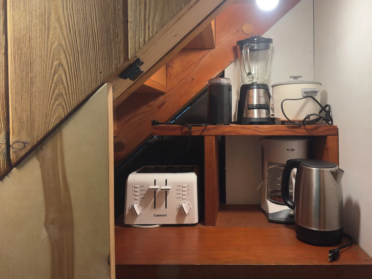 Small kitchen appliances including a toaster, blender, electric kettle, and coffee maker arranged on wooden shelves beneath a staircase.