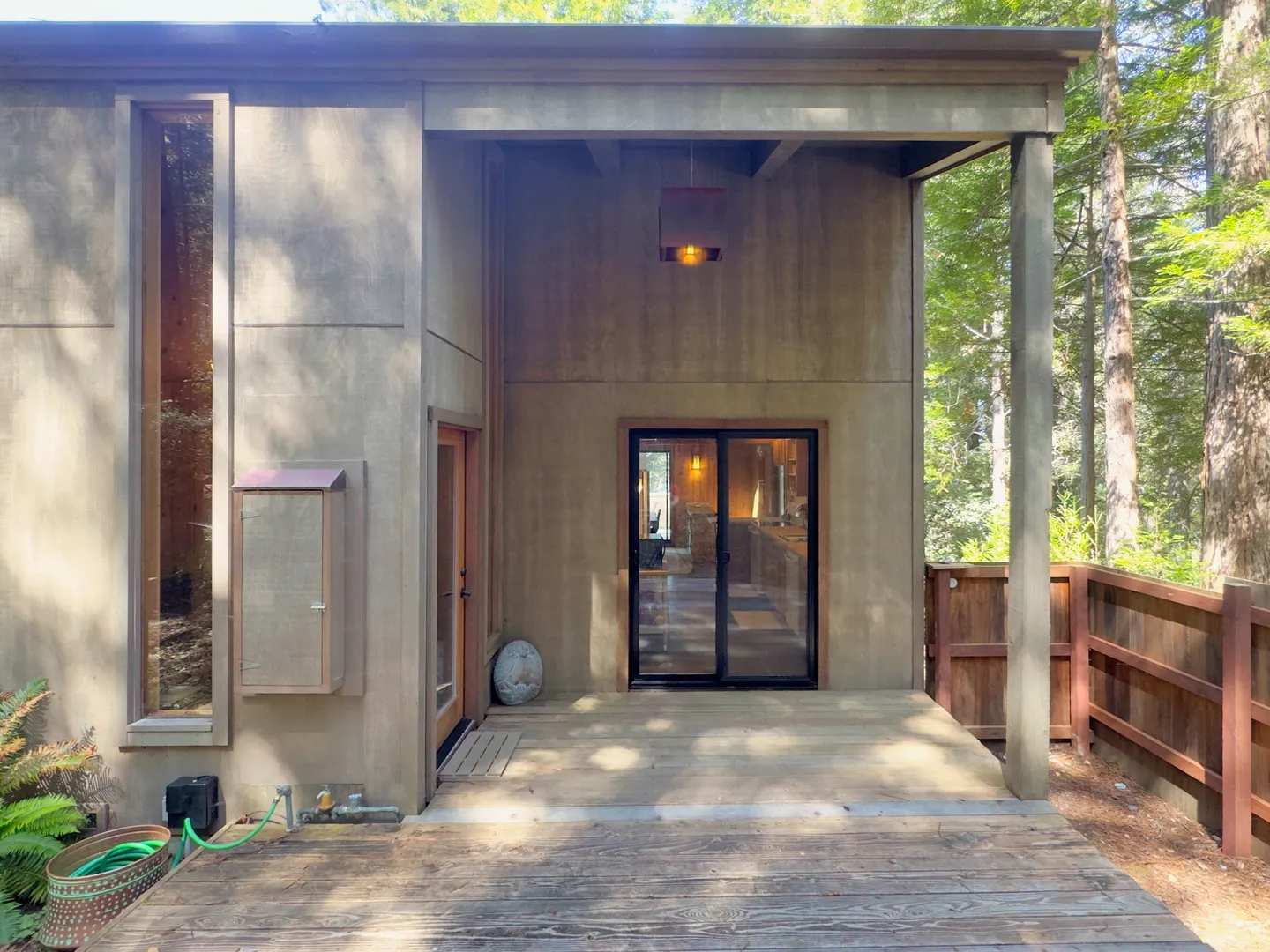 Exterior covered back porch with wooden deck, glass sliding door, and tall vertical window set in a wooded environment.