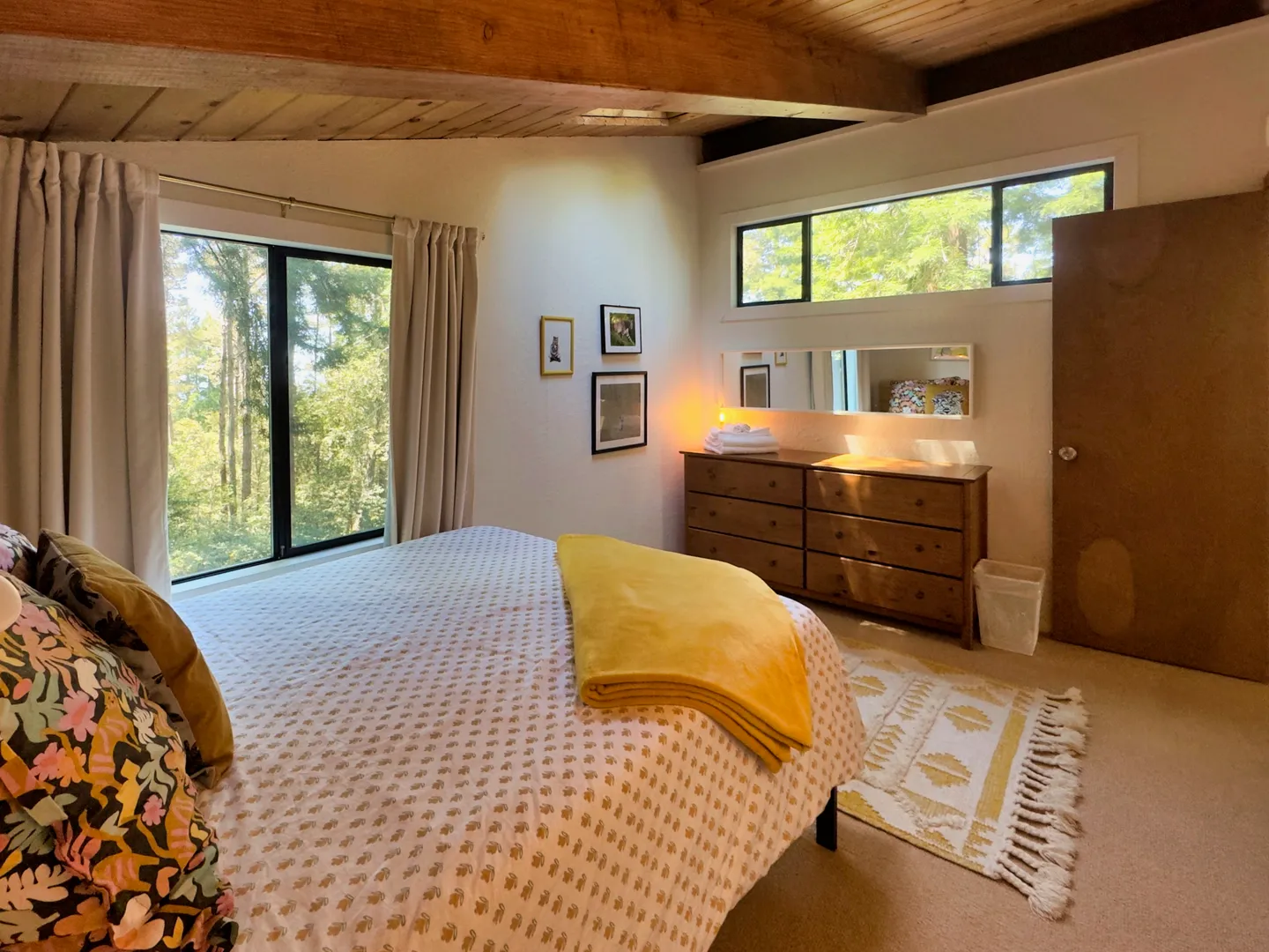 Bedroom with wood-beamed ceiling, large windows showing trees, a bed with a patterned duvet, and a wooden dresser.