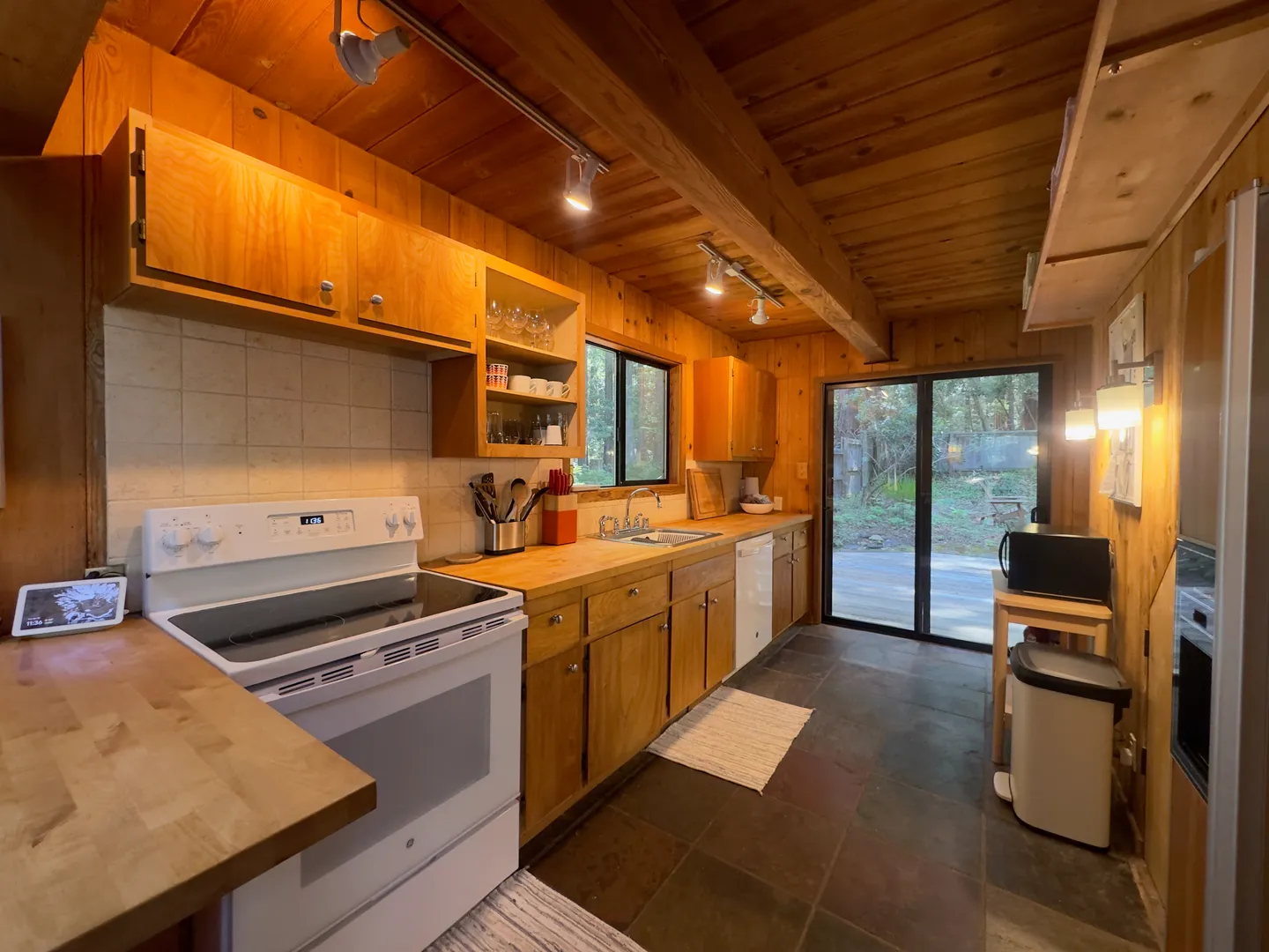 Rustic kitchen with wood cabinetry, wood-paneled walls and ceiling, white appliances, and a sliding glass door leading outside.