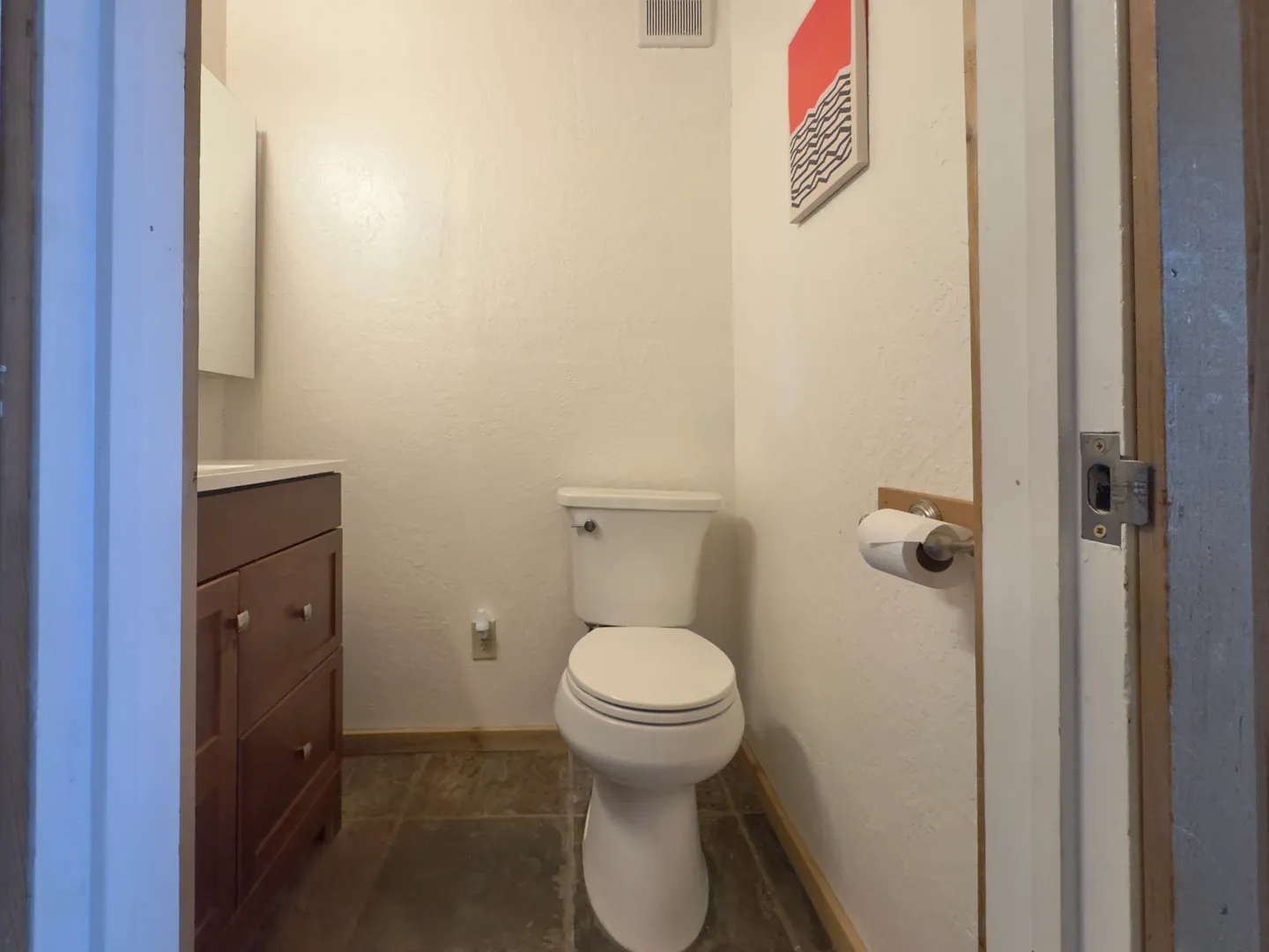 Bathroom viewed through an open doorway, featuring a white toilet, brown wood vanity, and tiled floor.