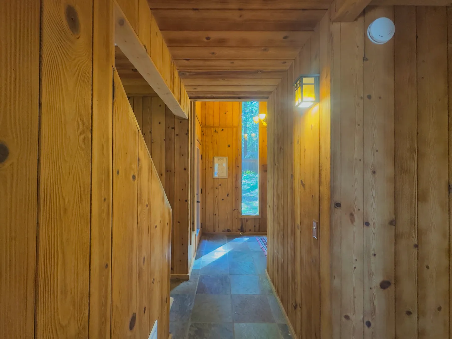 Wood-paneled hallway with tile flooring, a wall sconce, and a tall, narrow window at the end.