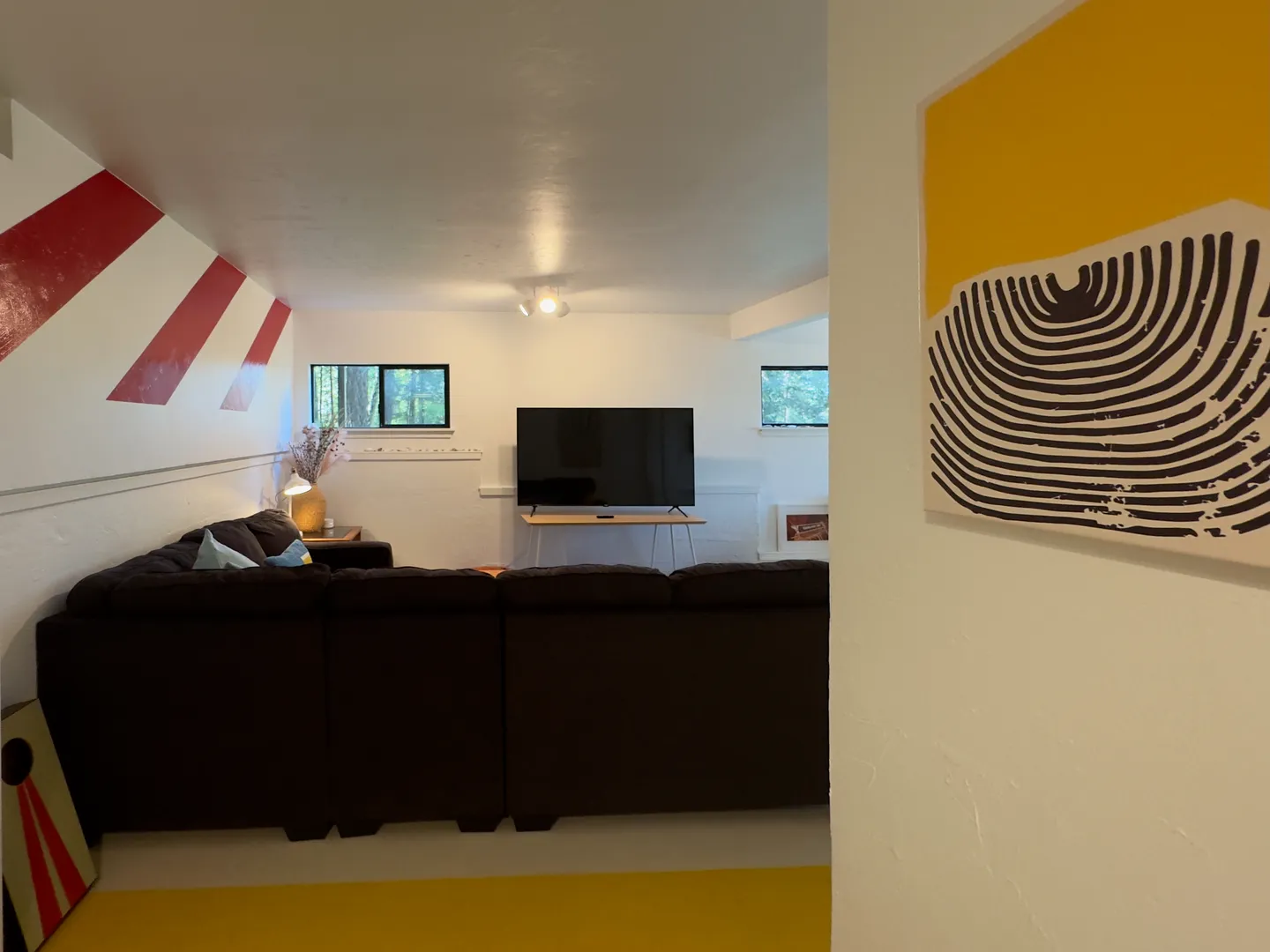 A living room with a large dark sectional sofa facing a flat-screen TV and white walls with red and white striped wall art.