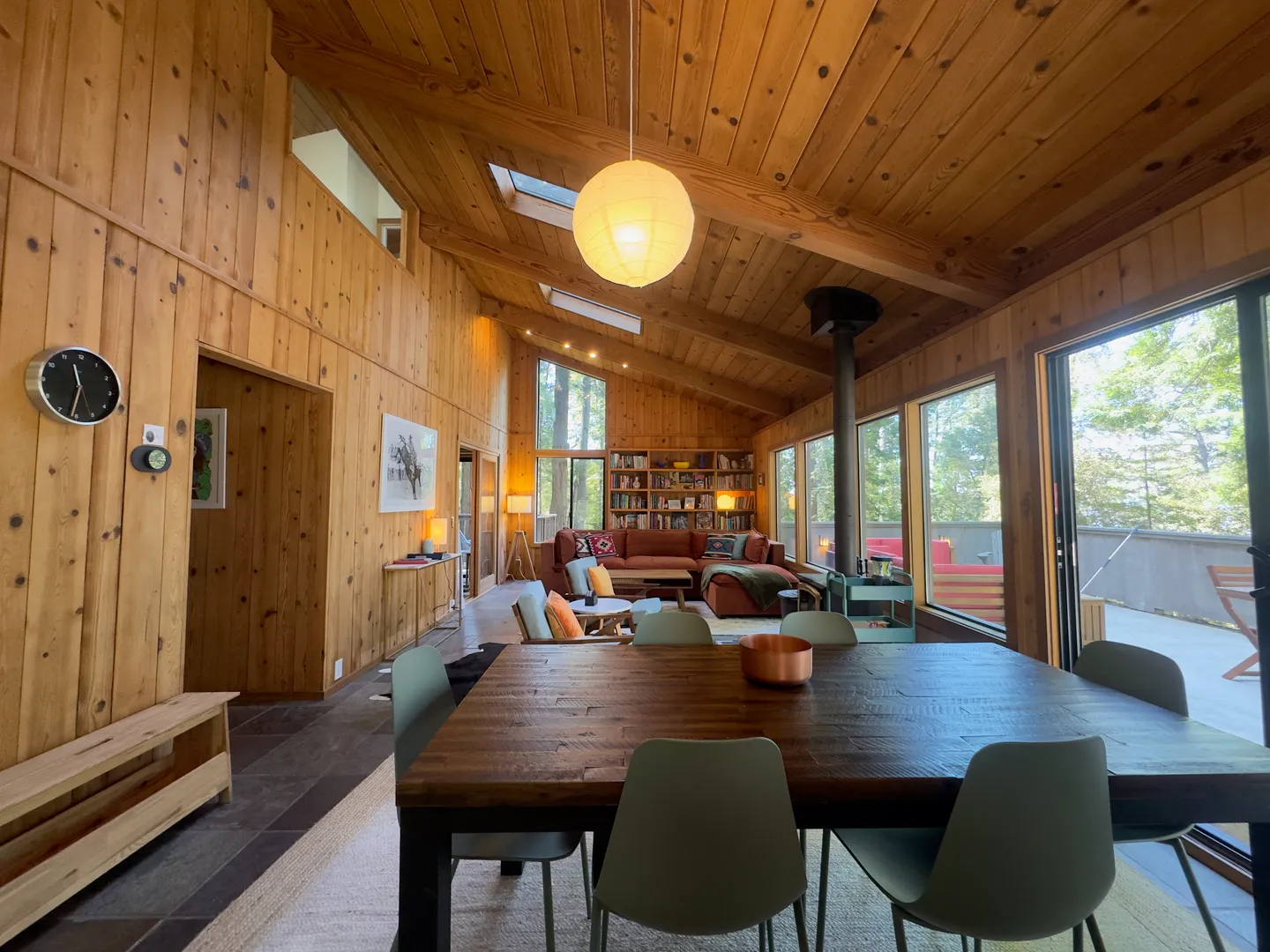 Open-concept dining and living room with wood-paneled walls and ceiling, skylights, dining table, and large windows looking outside.