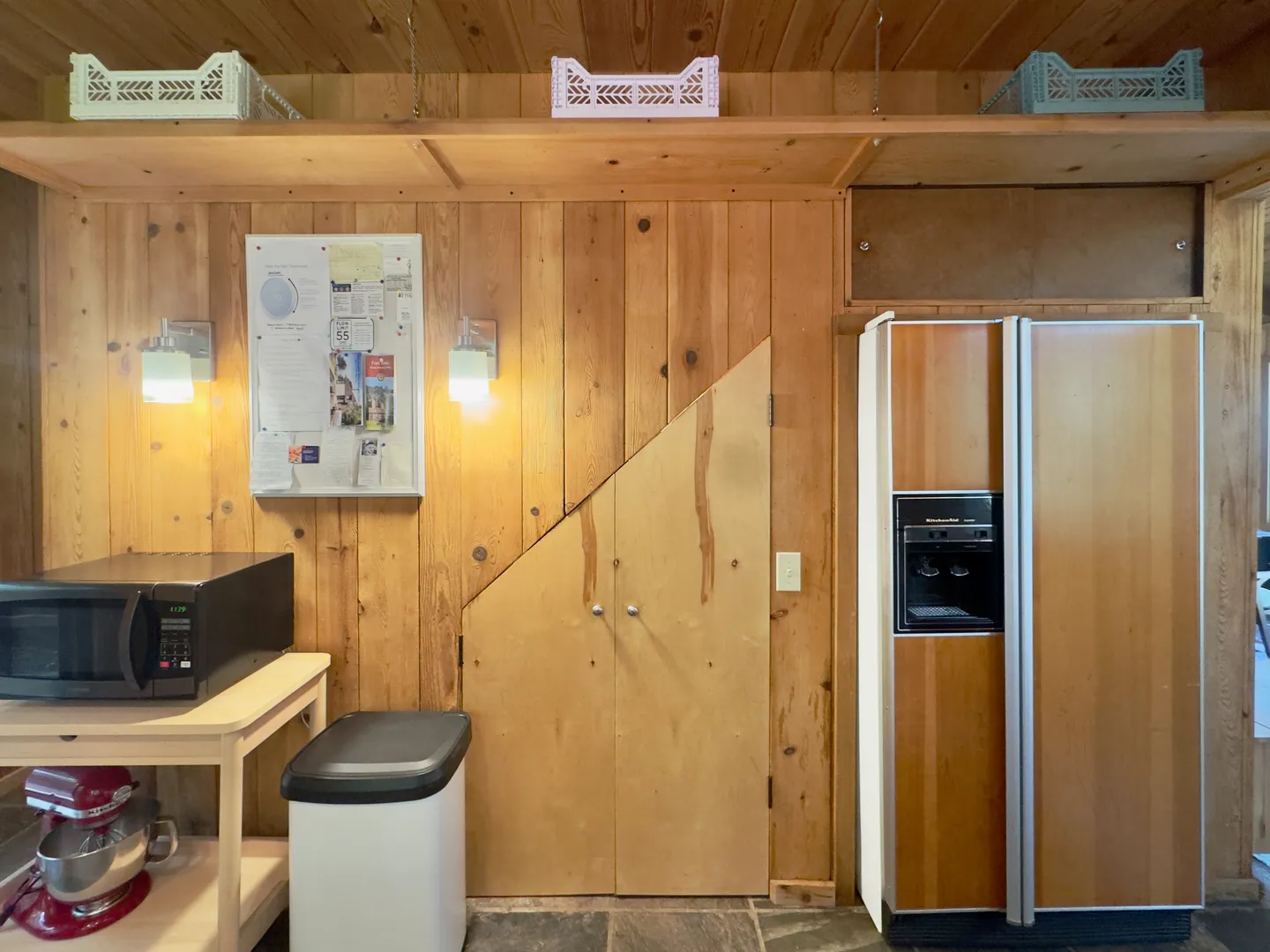 A kitchen nook with pine wood paneling, an angled cabinet door, a refrigerator with wood-paneled front, a microwave on a rolling cart, and an upper storage shelf.
