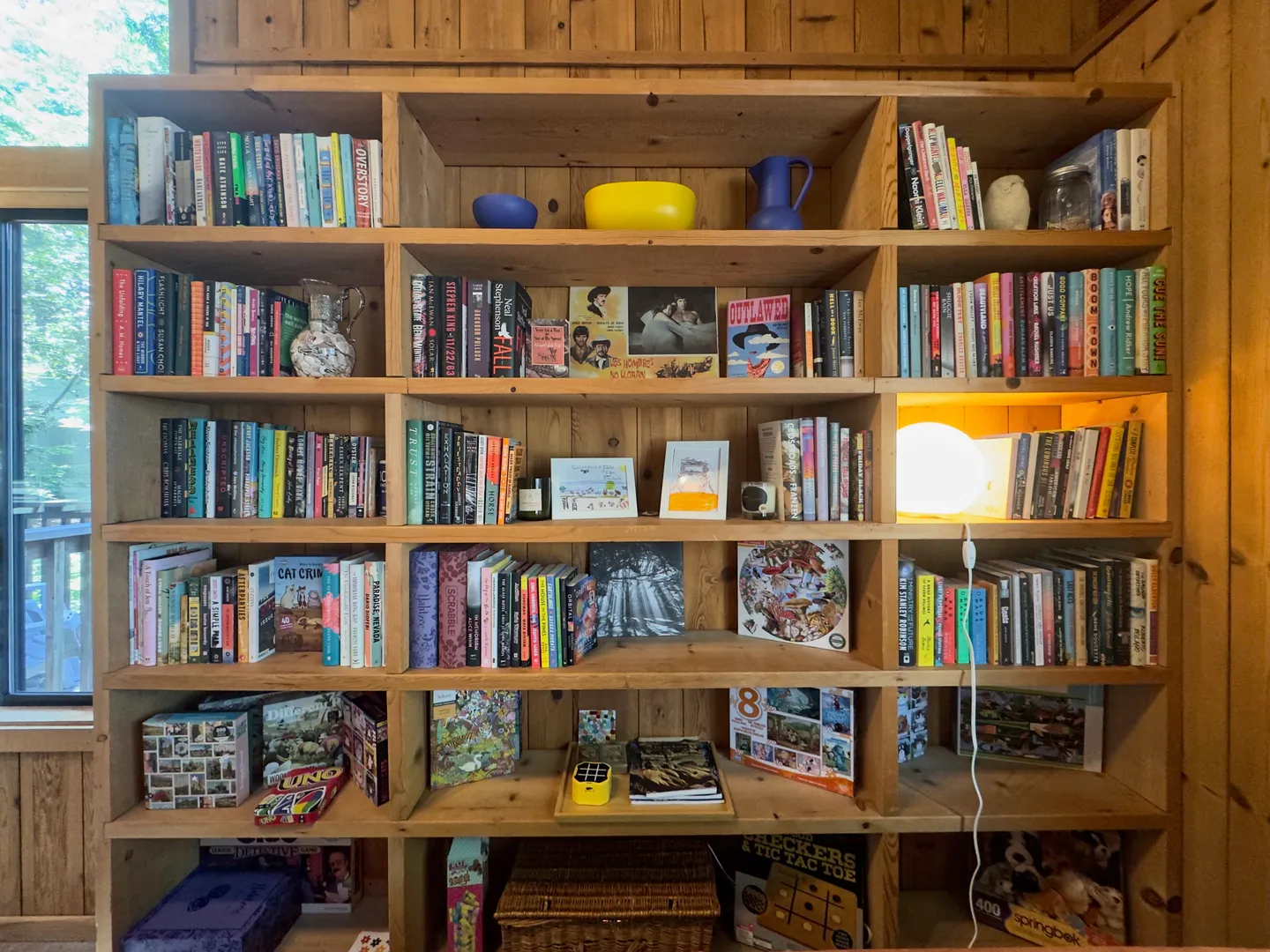 Large built-in light wood bookcase filled with numerous books, decorative bowls, and board games against a wood-paneled wall.