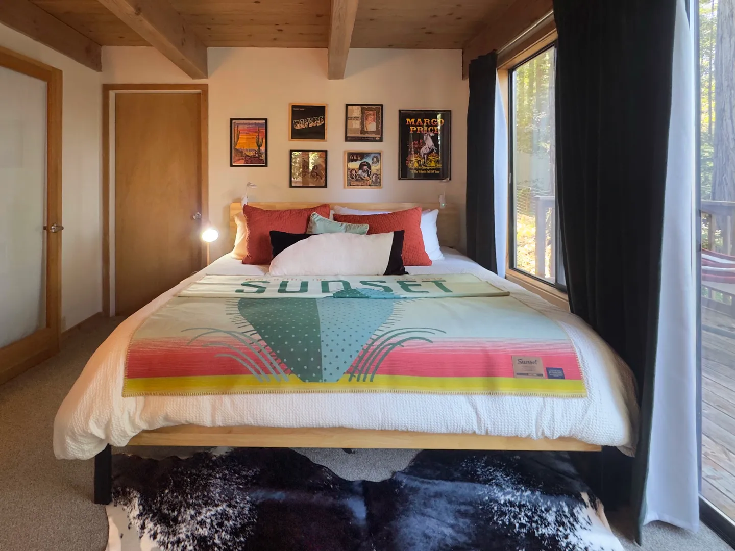 Bedroom with a wood-beam ceiling, bed styled with a colorful patterned blanket, framed wall art, and large sliding glass doors.