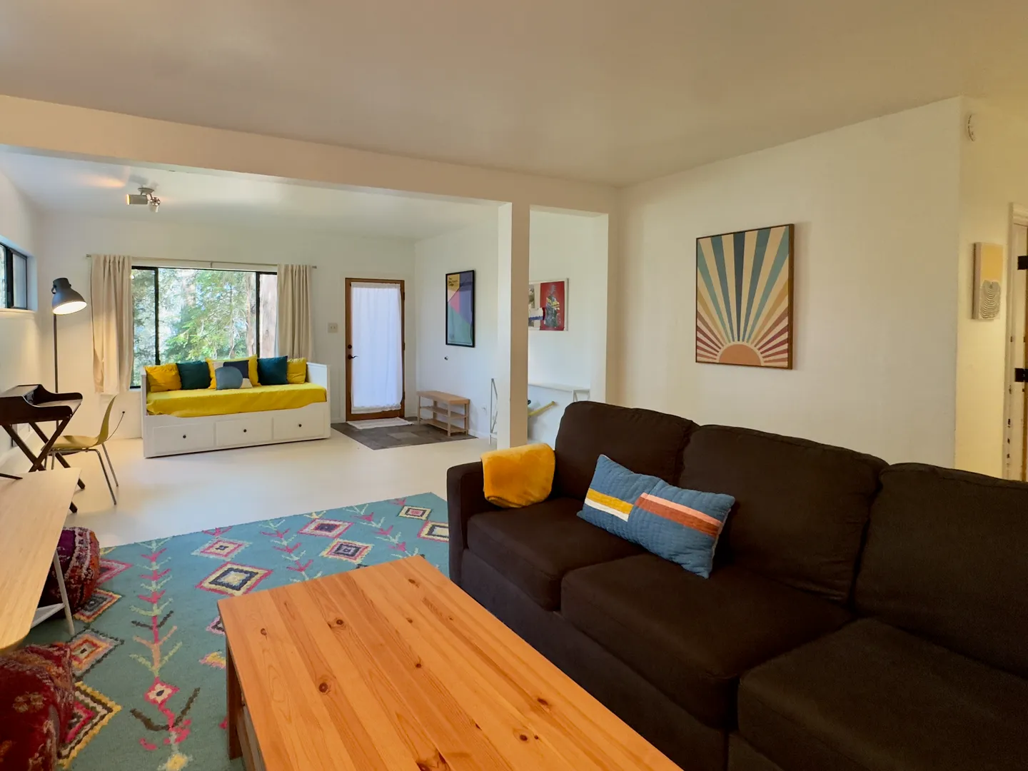 Living area with a dark sectional sofa, wood coffee table, patterned teal rug, and an adjoining open room featuring a daybed and an exterior door.