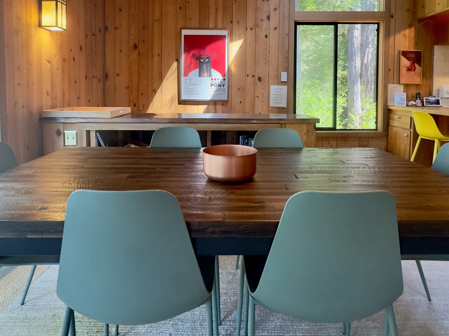 Dining area featuring a dark wood plank dining table surrounded by pale green modern chairs in a room with wood-paneled walls.