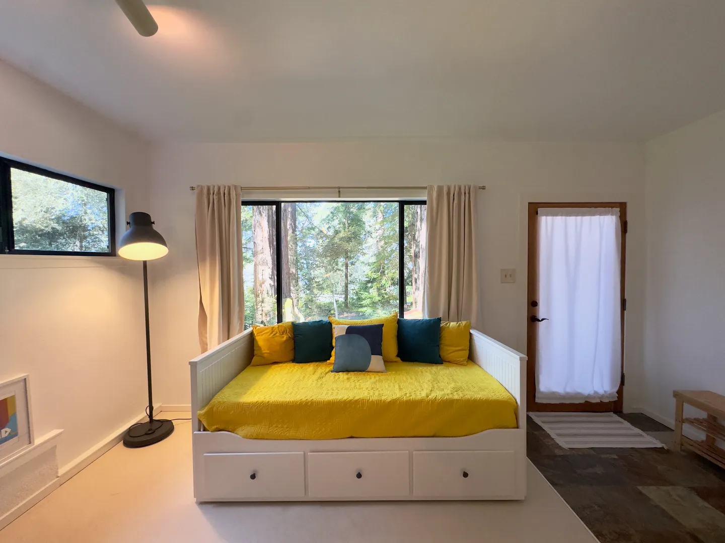 A white daybed with a yellow cover and colorful accent pillows sits beneath a window overlooking wooded scenery.