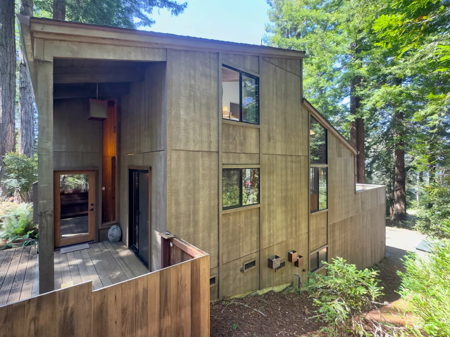 Exterior of a modern multi-level wood-sided home surrounded by tall redwood trees with a wooden deck and entrance.