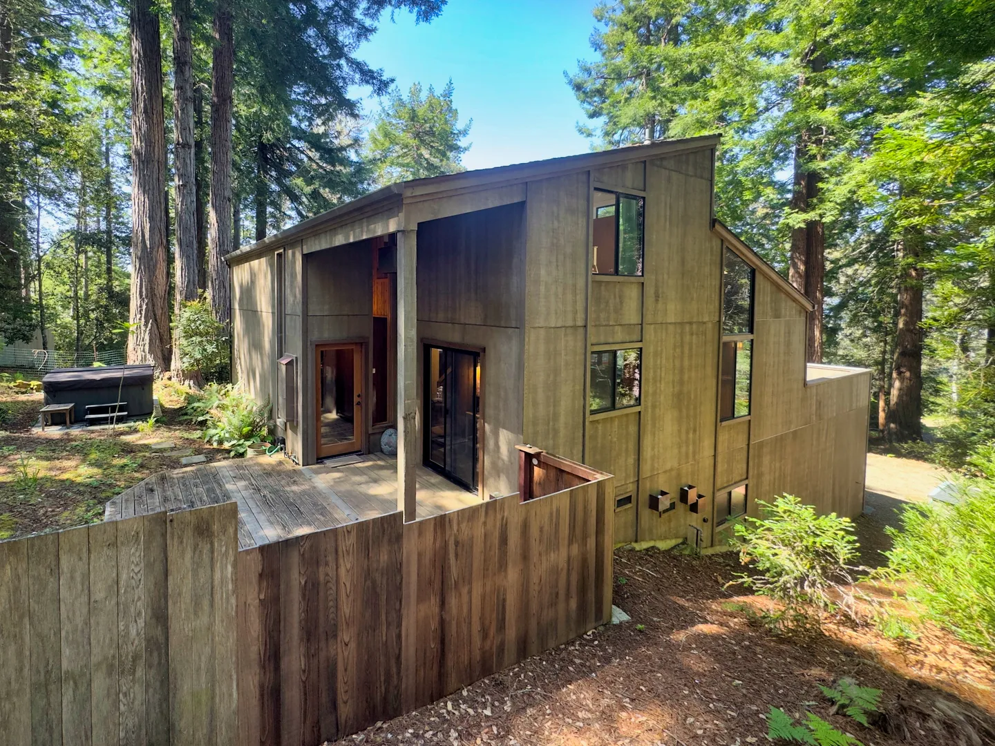 Exterior view of a multi-level wood-sided cabin in the woods with a deck and hot tub.