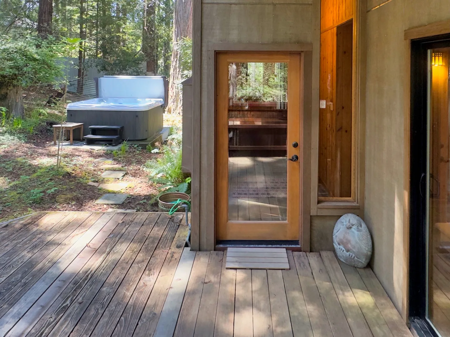 A wooden outdoor deck leading to a glass-paneled door, with an outdoor hot tub situated in the wooded yard nearby.
