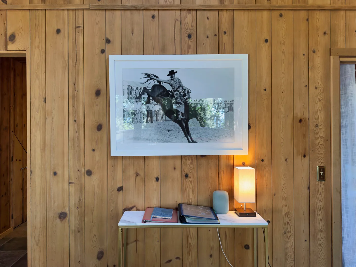 A framed black-and-white rodeo photograph hung above a marble-topped console table against vertical knotty pine wood paneling.