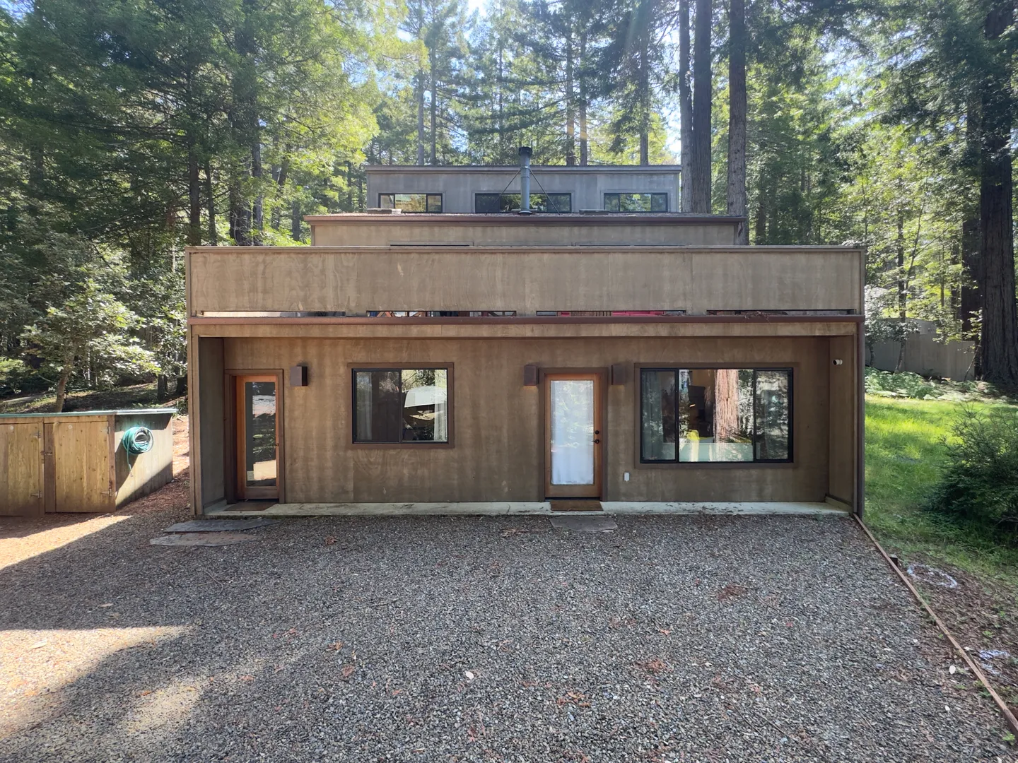 Exterior view of a multi-level wood-sided house surrounded by tall pine trees, featuring a gravel driveway in front.