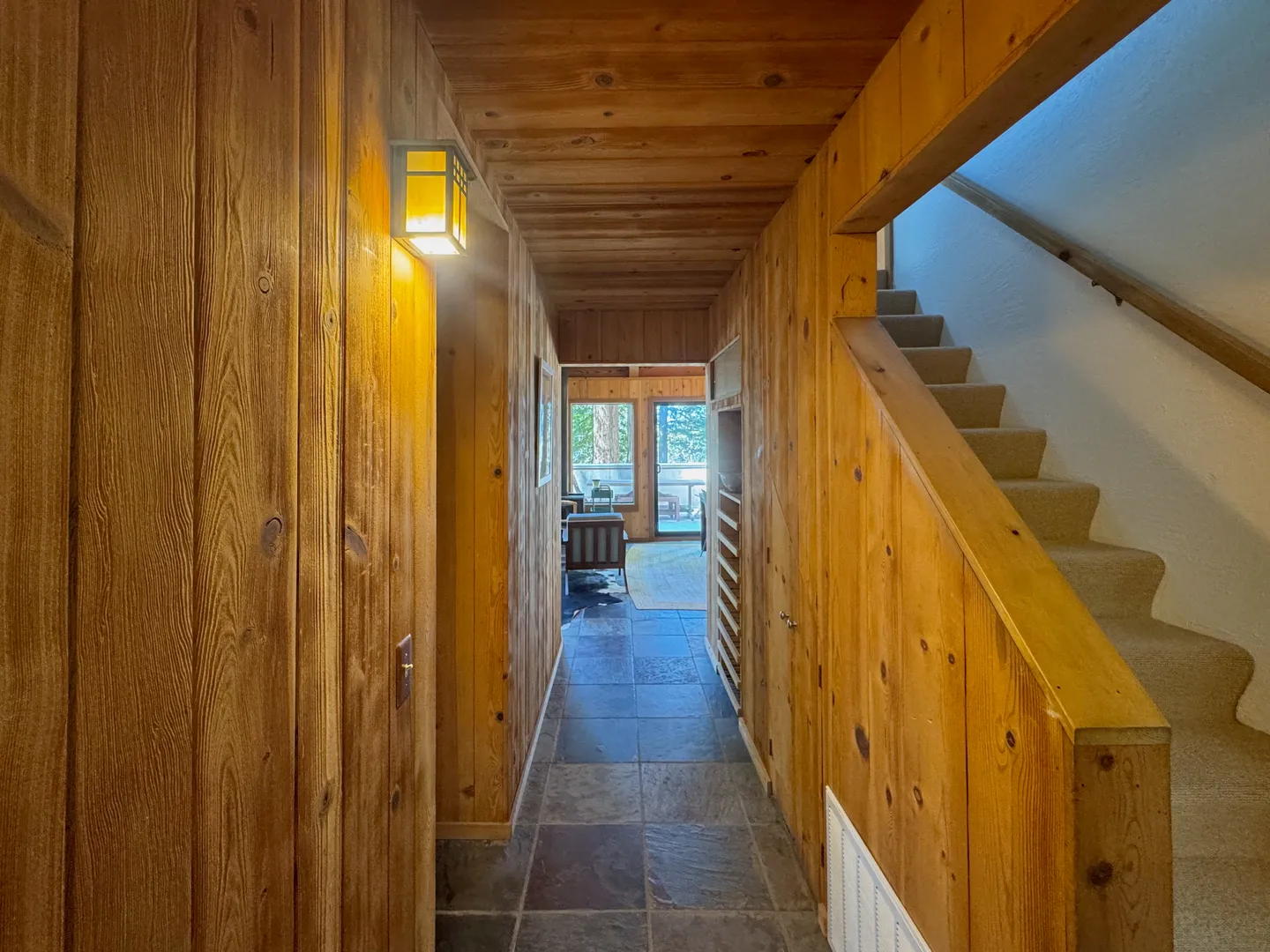 Hallway lined with vertical wood paneling and slate-colored tile flooring, with a carpeted staircase to the right.