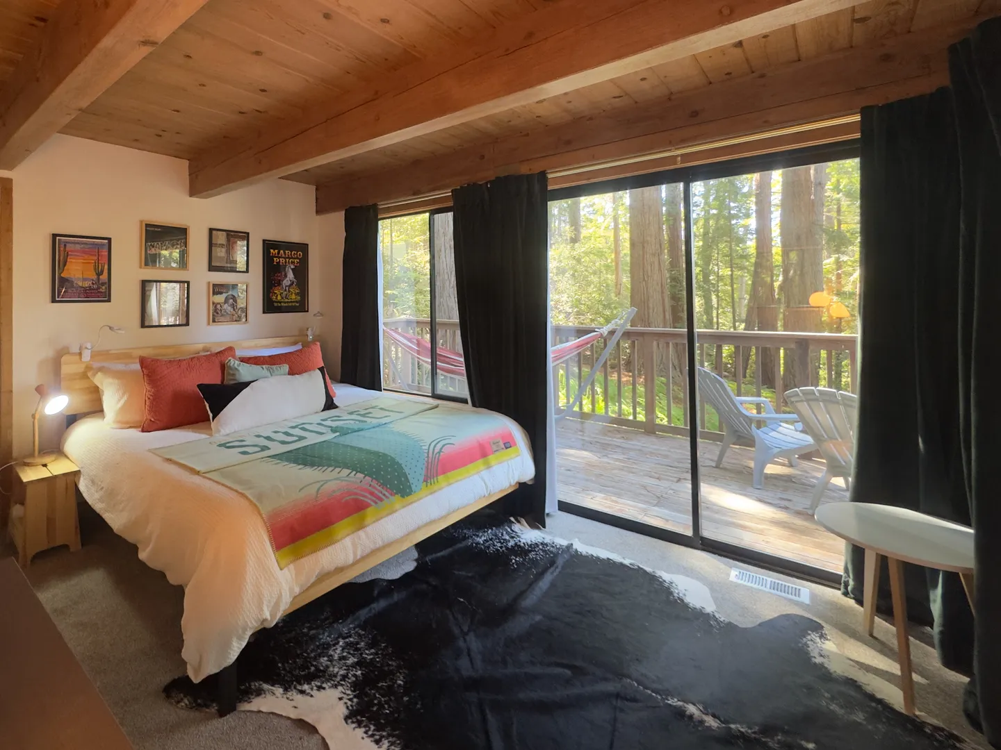 Bedroom with wooden ceiling beams, a bed with colorful accents, and large glass sliding doors opening to a forest-view deck.