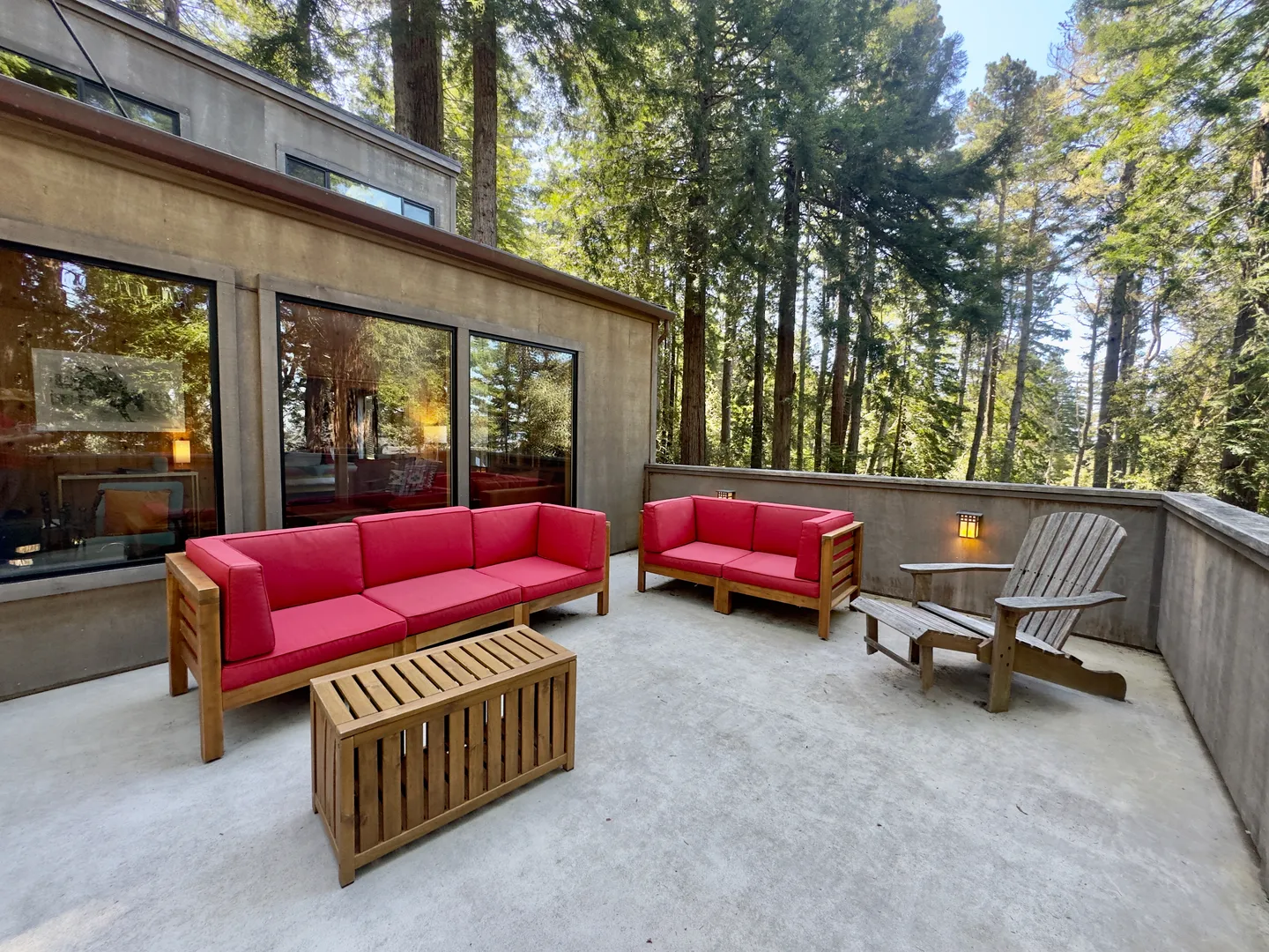 Outdoor patio with concrete flooring, red-cushioned wooden seating, a slatted coffee table, and an Adirondack chair bordered by dense evergreen trees.