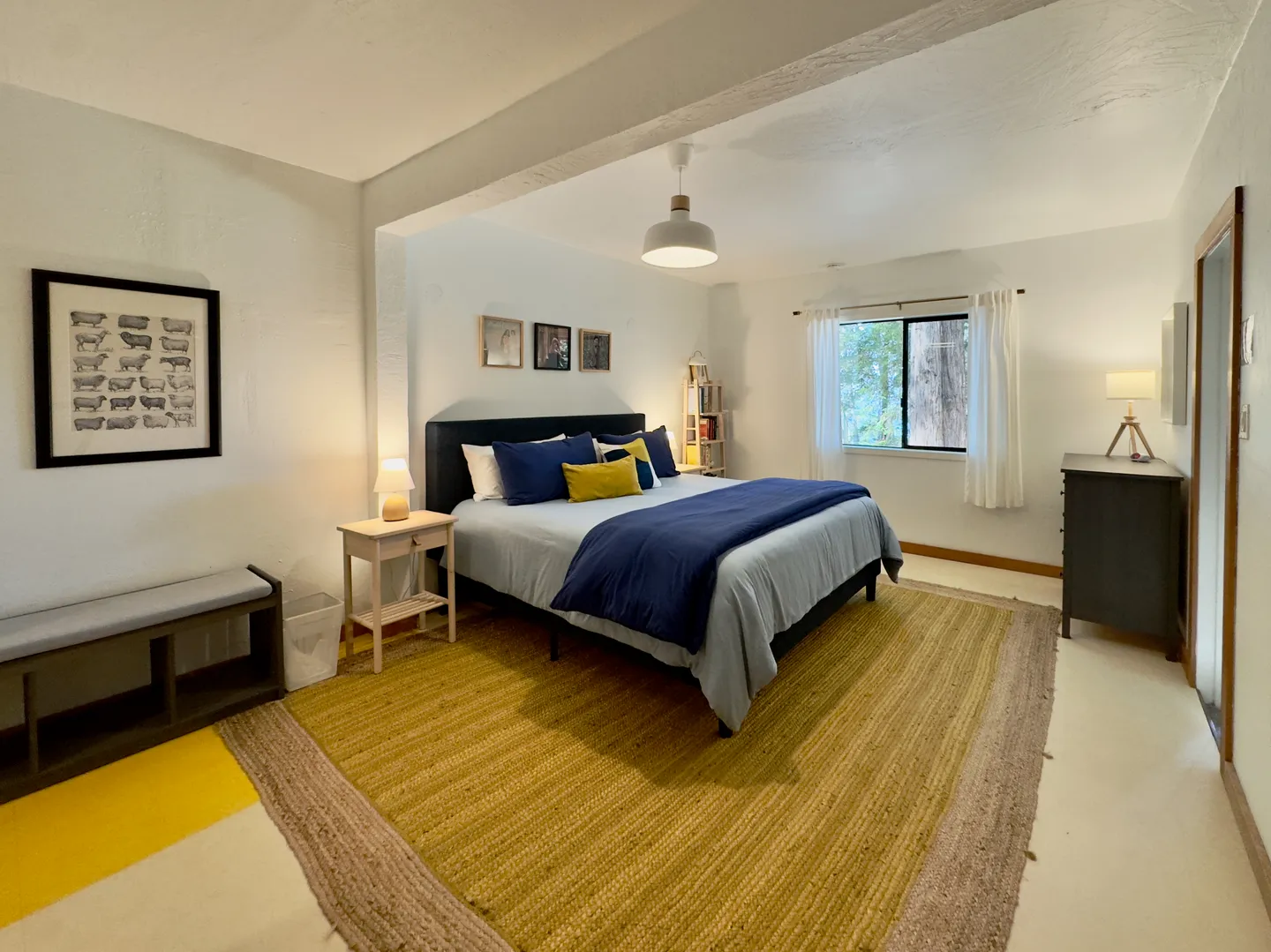 Bedroom featuring a made bed with navy blue and yellow accents, an area rug, nightstands, and a window with white curtains.