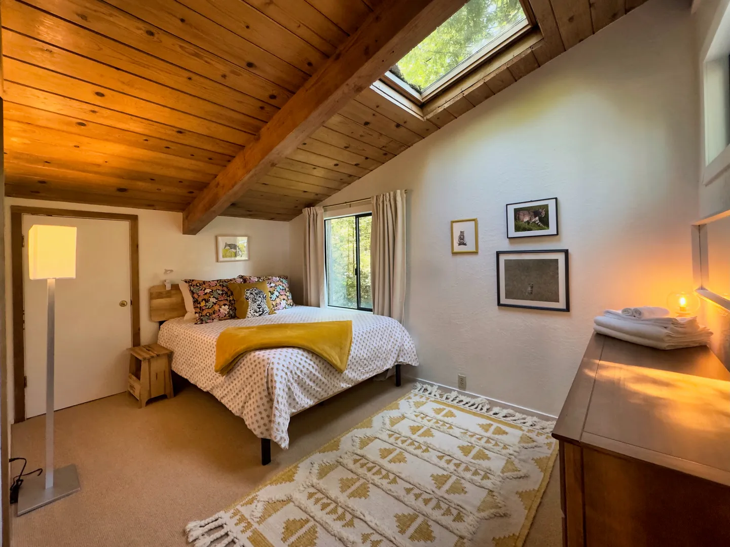 Bedroom with vaulted wood-paneled ceiling, skylight, a made bed, patterned rug, dresser, and window.