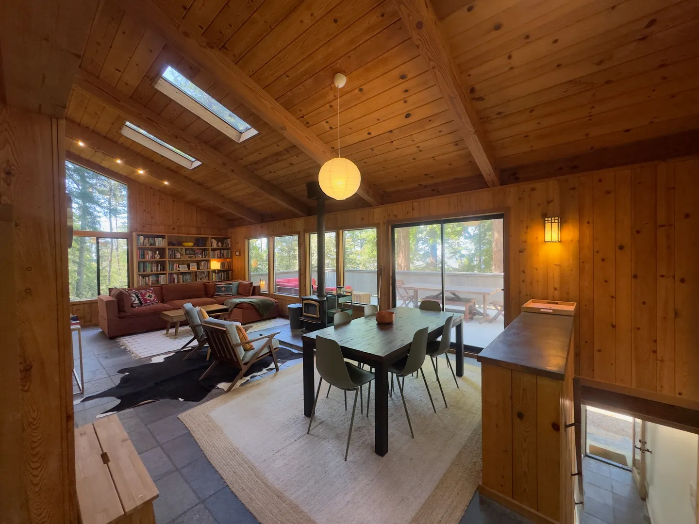Open-concept cabin interior with wood-paneled walls and ceiling, dining area, and living room with sectional sofa and wood stove.