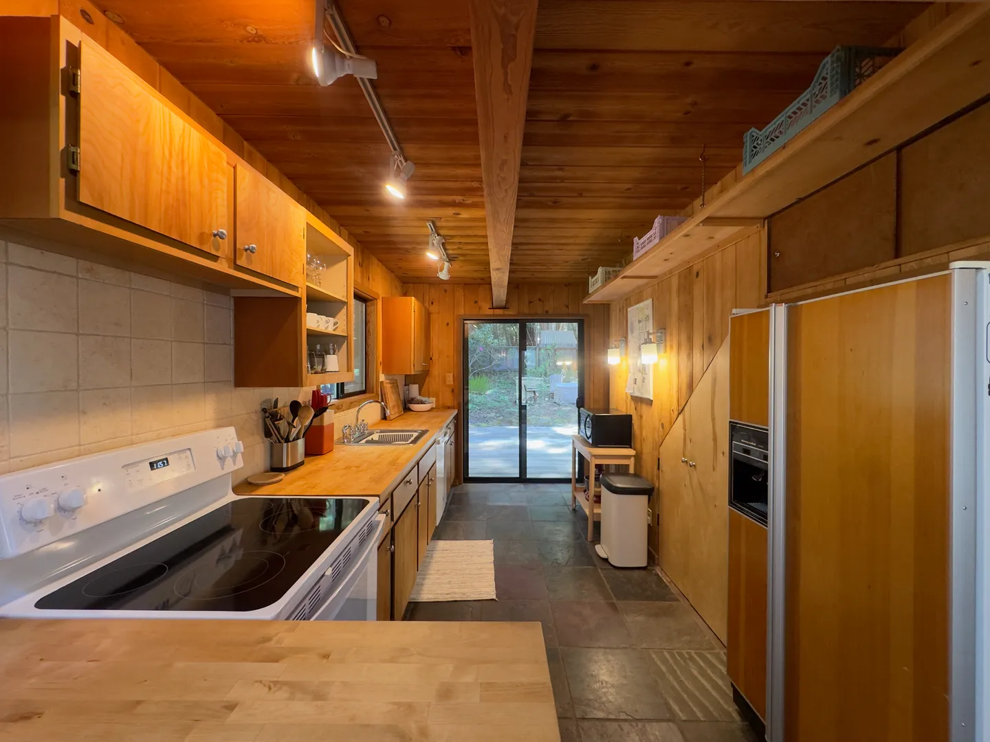 Galley-style kitchen with natural wood walls, wood cabinets, a white electric range, and glass sliding doors leading outdoors.