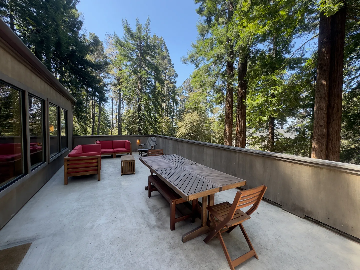 Outdoor patio with a wooden dining table, bench, chair, and a red-cushioned sectional sofa surrounded by tall evergreen trees.