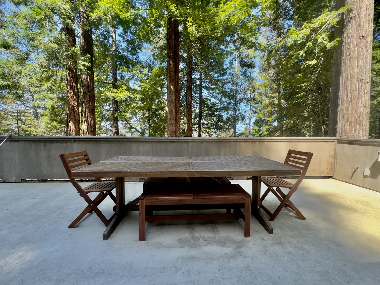 An outdoor dining table with wooden chairs and benches situated on a concrete patio bordered by tall trees.