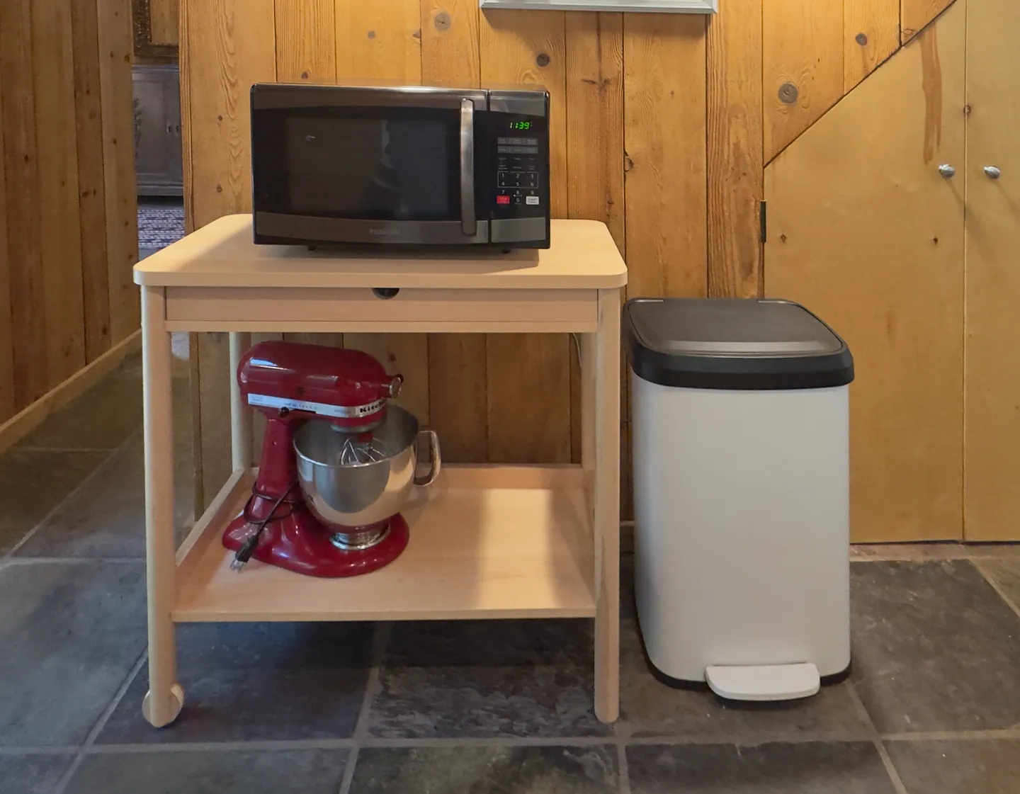 A small wooden cart with a microwave on top and a red stand mixer below, positioned next to a white trash can against a wood-paneled wall.