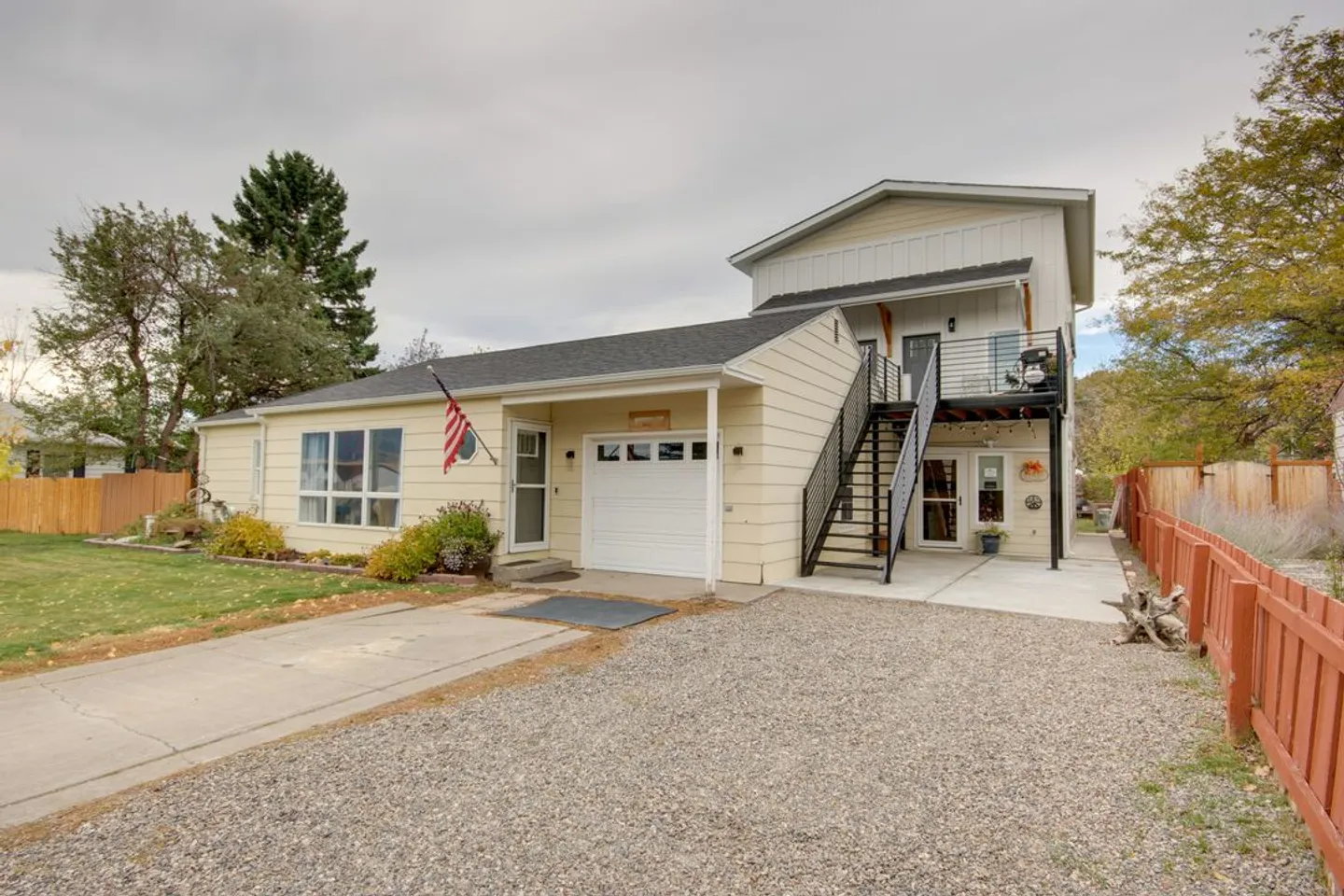Exterior view of a multi-level home with pale yellow siding, an attached garage, an exterior metal staircase, and a gravel driveway.
