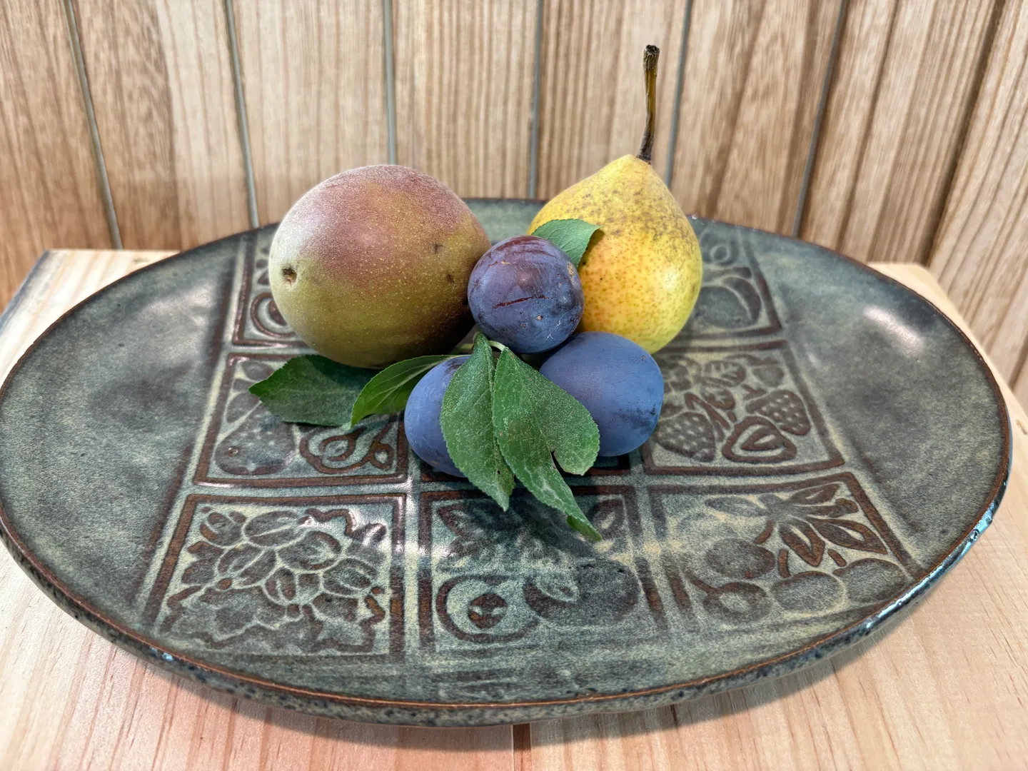 A ceramic plate featuring embossed patterns holding fresh fruit including a pear and plums against a wood-paneled background.