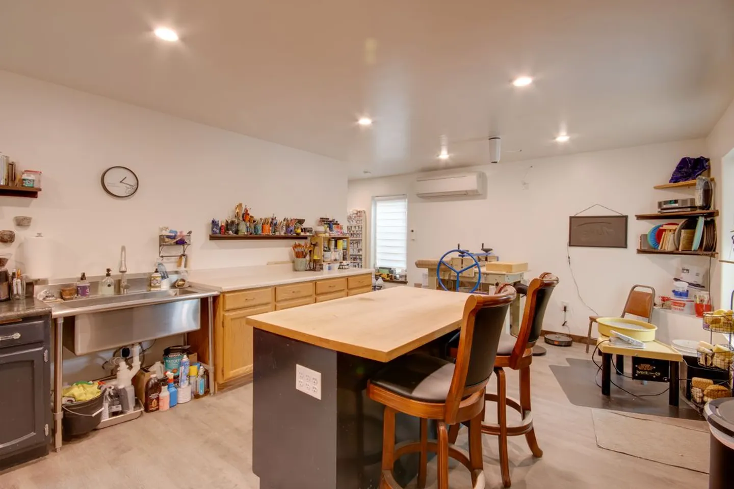 An art or pottery studio featuring a central work island with barstools, a stainless steel utility sink, countertops, and pottery equipment.