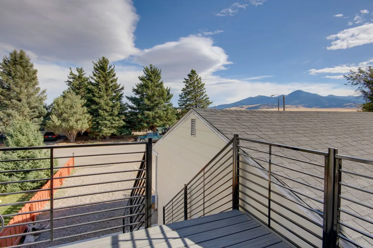 View from an elevated exterior deck with black metal horizontal railings looking over a rooftop toward pine trees and distant mountains under a blue sky.