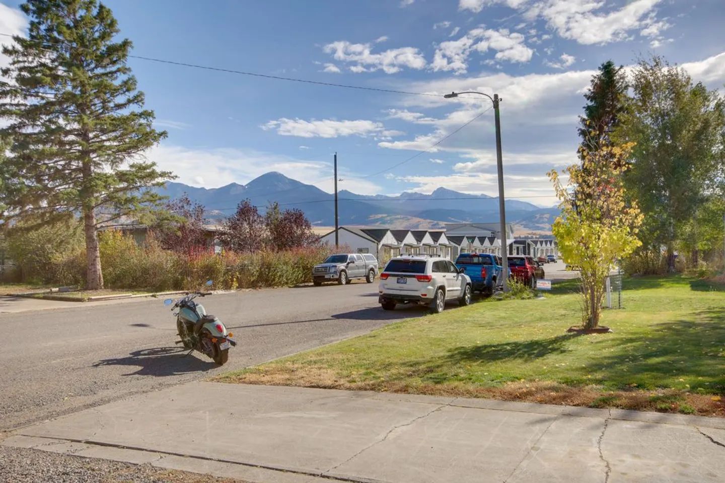 Street view of a neighborhood road with parked cars, a motorcycle, grassy lawns, and mountains in the distance under a blue sky.