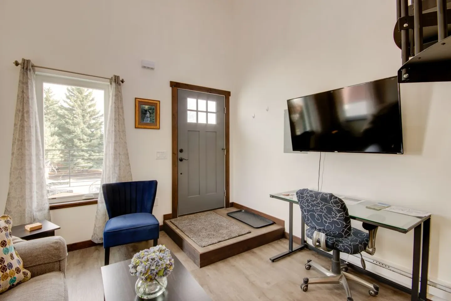 Living area with entry door, blue armchair, window, and a small desk with a patterned office chair beneath a wall-mounted TV.