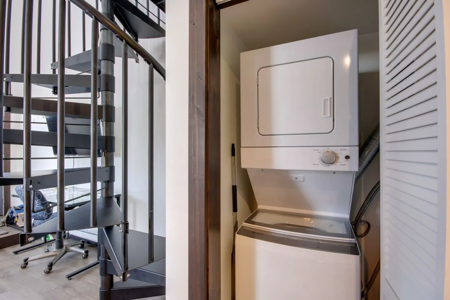 A stacked washer and dryer in a closet adjacent to a black metal spiral staircase.