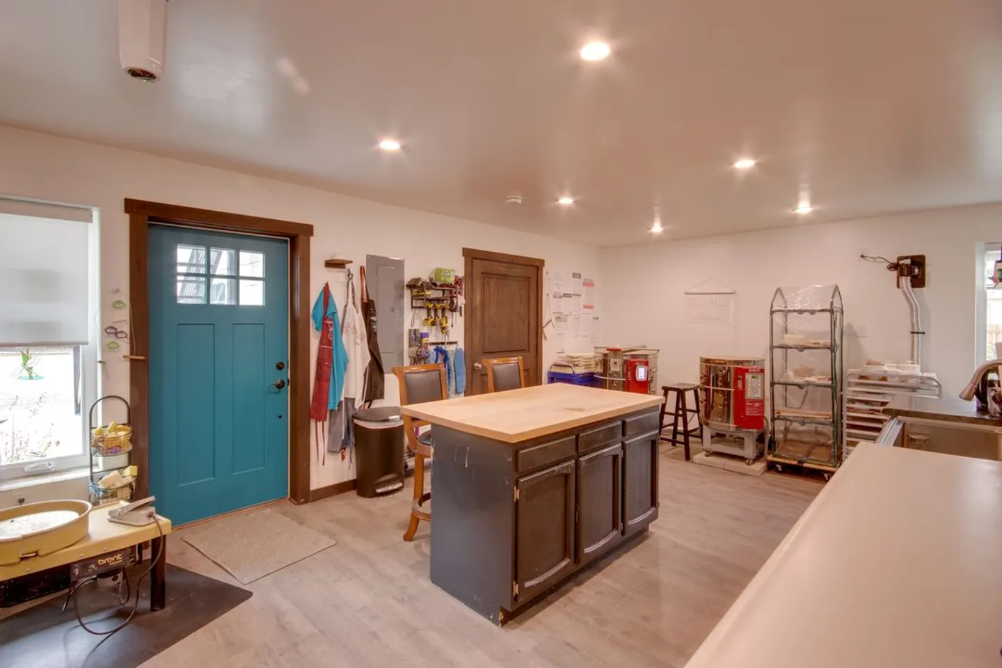 A pottery or craft studio room with a center wooden-topped island, pottery wheel, kiln, shelving, and a bright blue exterior door.