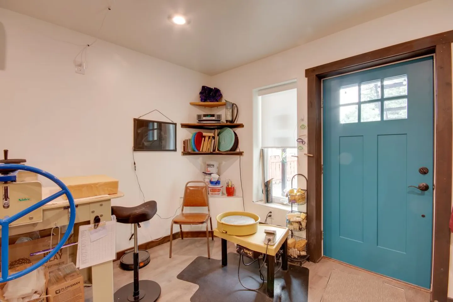 A pottery studio workspace featuring a pottery wheel, tools, work table, and a bright blue exterior door.