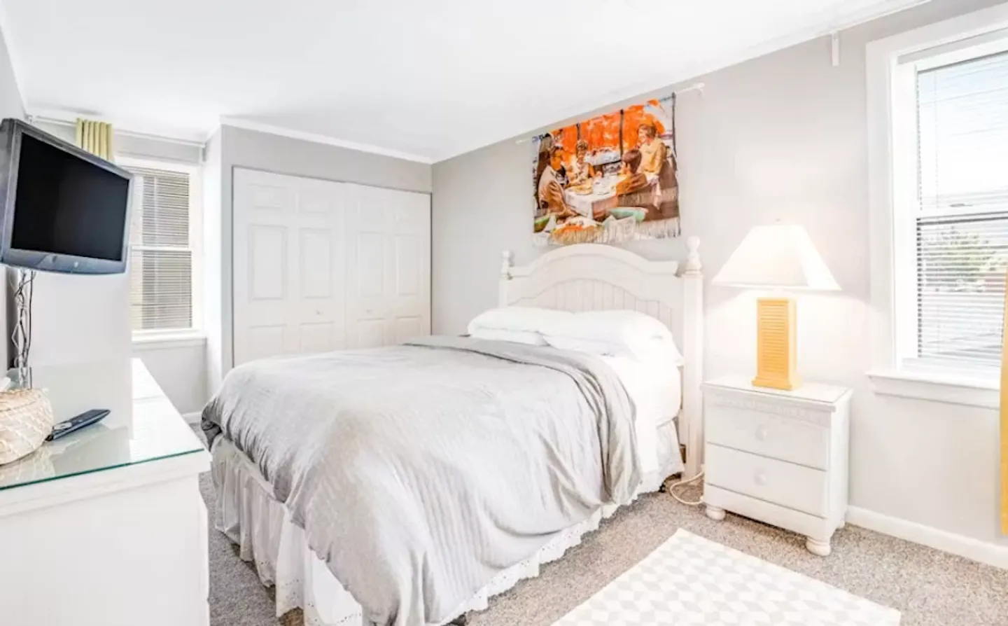 Bedroom with a white headboard bed, nightstand with lamp, dresser with mounted TV, and closet doors.