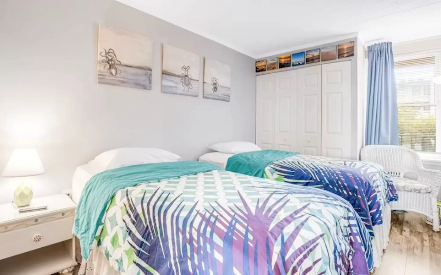 Bedroom with two twin beds dressed in patterned blue and green palm leaf bedding, a white nightstand, and bifold closet doors.