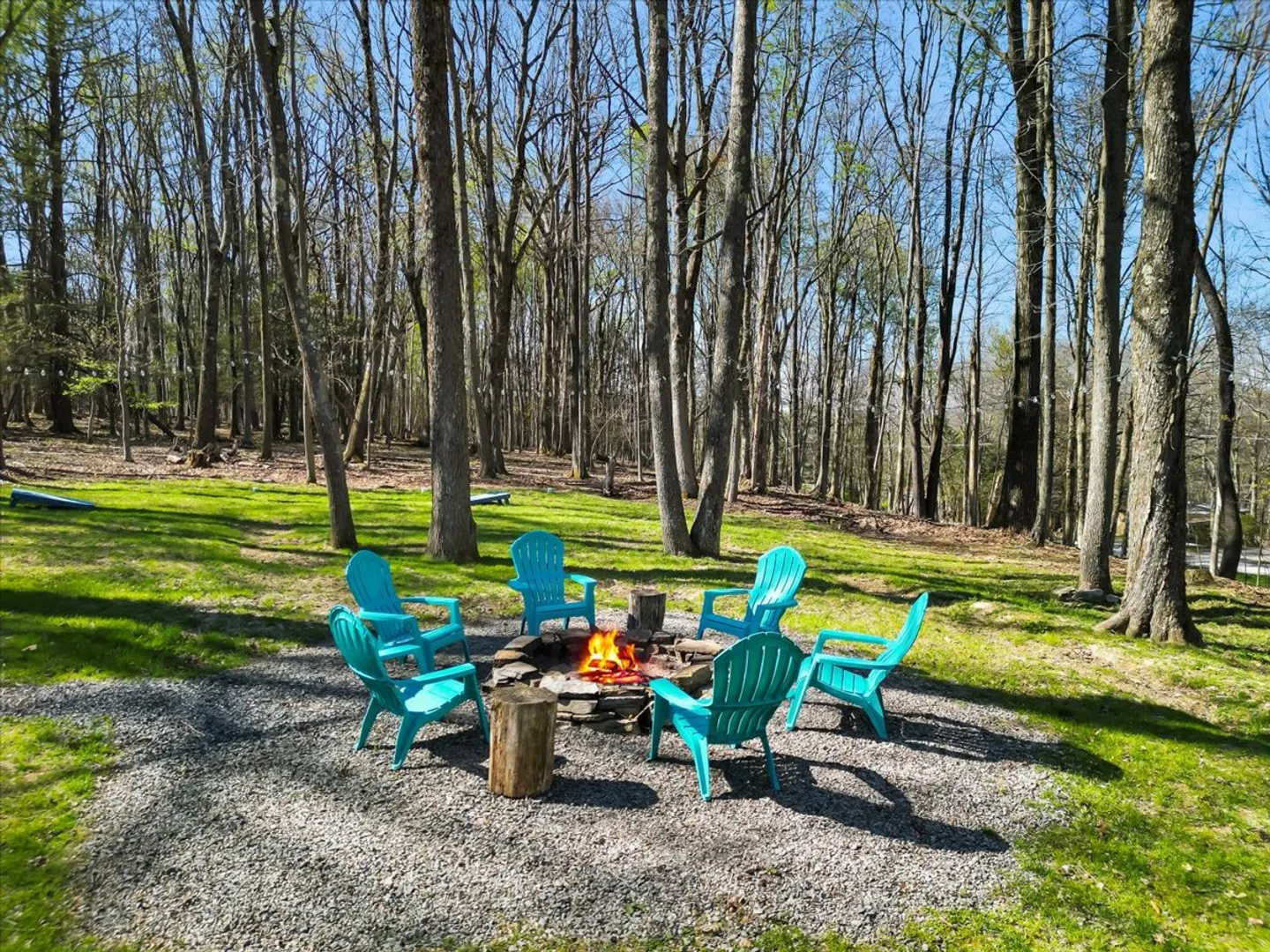 Outdoor gravel fire pit area with a burning fire surrounded by turquoise Adirondack chairs, log side tables, and wooded surroundings.